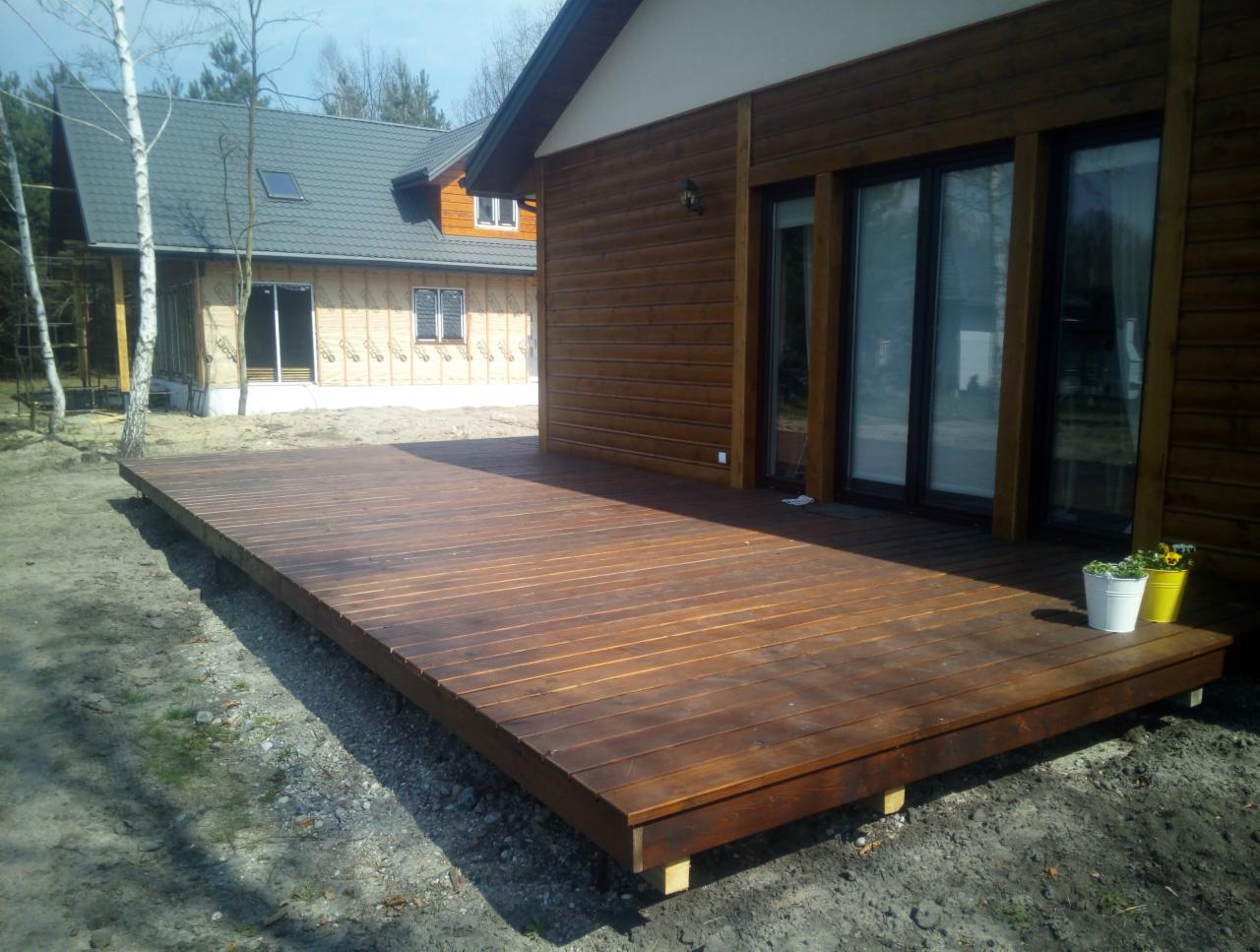 Rozległy drewniany taras przylegający do domu z drewnianą elewacją, w tle drugi dom w trakcie budowy, doniczki z kwiatami na tarasie.