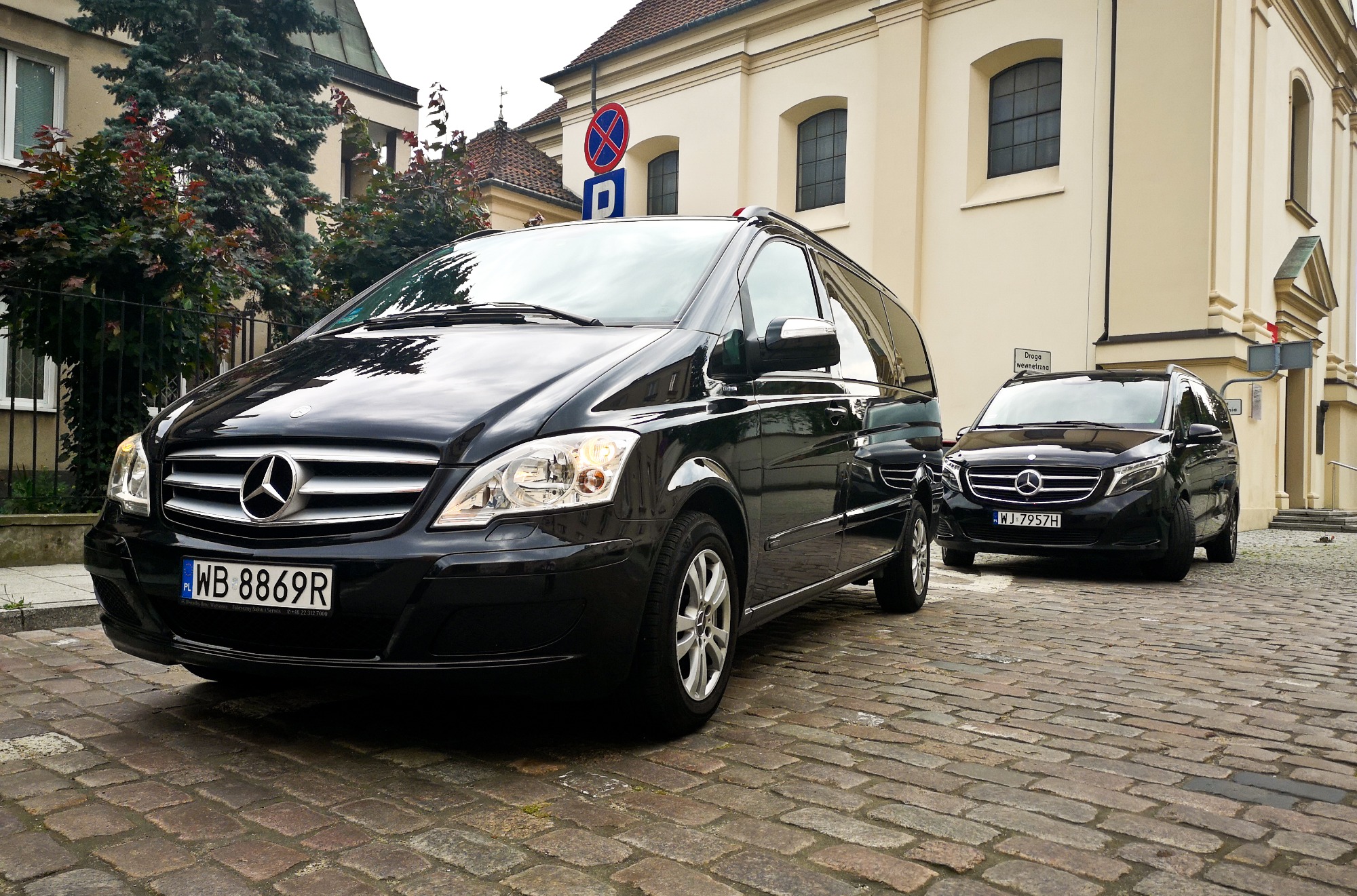 Dwa czarne busy Mercedes-Benz V-klasy zaparkowane na brukowanej ulicy przed budynkiem z beżową elewacją, widoczny znak drogowy zakazu zatrzymywania się.