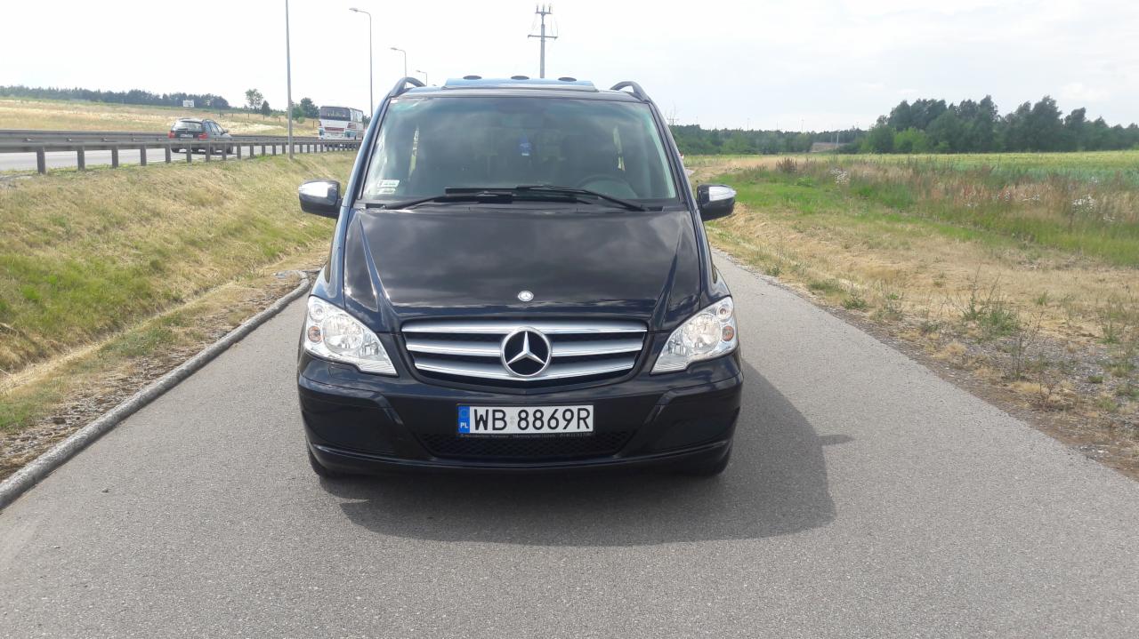 Czarny Mercedes Vito z widoczną tablicą rejestracyjną WB 8869R, zaparkowany na poboczu drogi asfaltowej z widokiem na autostradę i pola w tle.