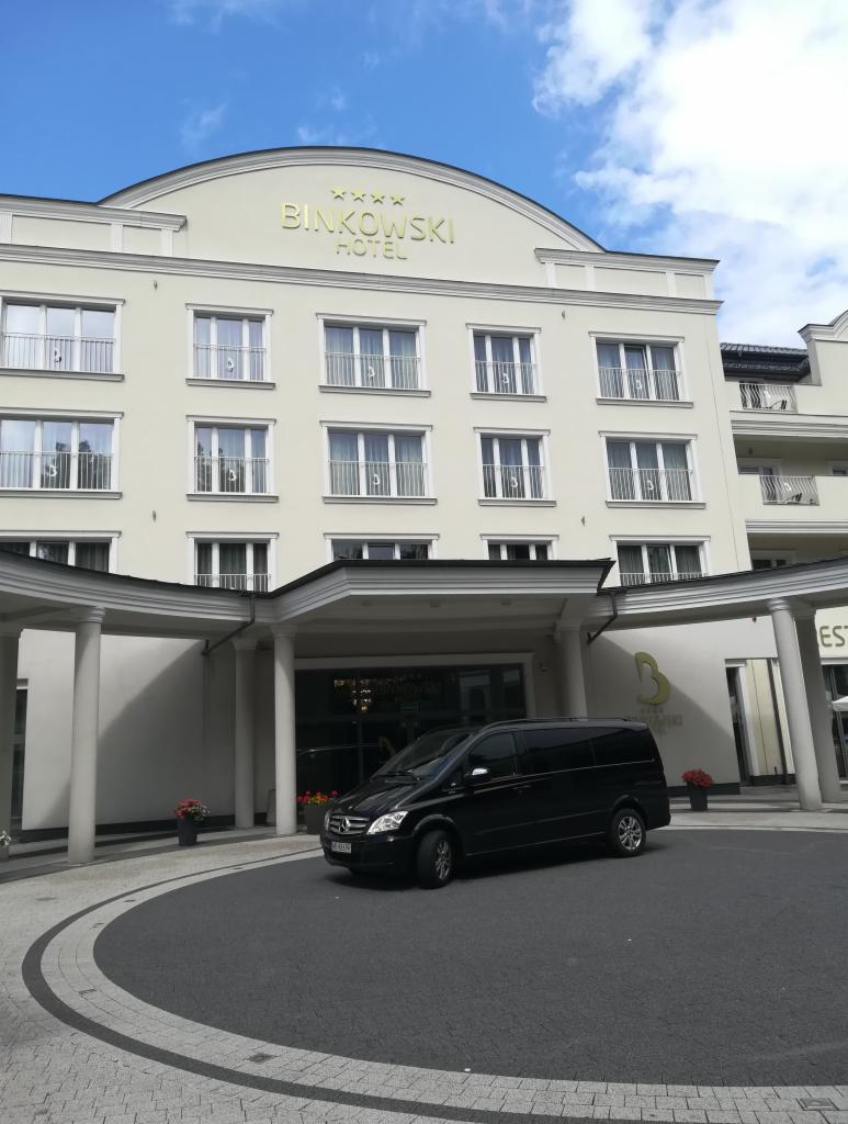 Czarny van Mercedes zaparkowany przed hotelem Binkowski z kolumnami i kwiatami w donicach.
