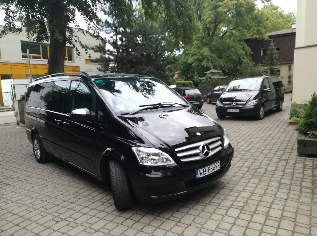 Czarny Mercedes-Benz Viano zaparkowany na brukowanym podwórku, widoczne tablice rejestracyjne i inne pojazdy w tle.