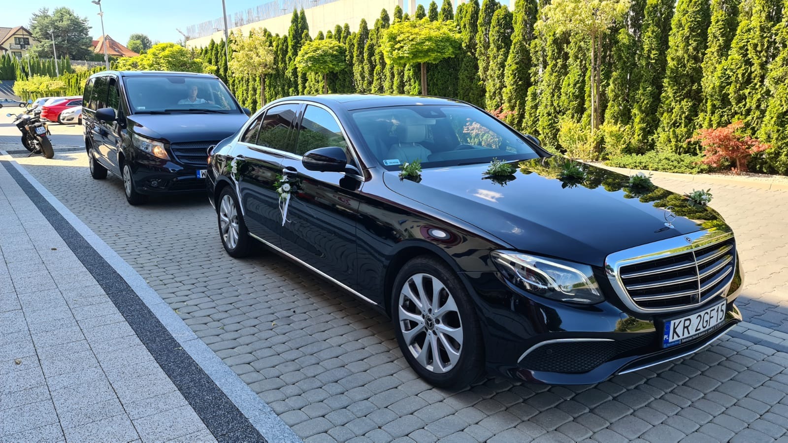 Dwa czarne, udekorowane kwiatami i wstążkami samochody Mercedes, jeden osobowy i jeden typu van, stoją na brukowanym podjeździe przed zielonym żywopłotem. Tablica rejestracyjna widoczna...