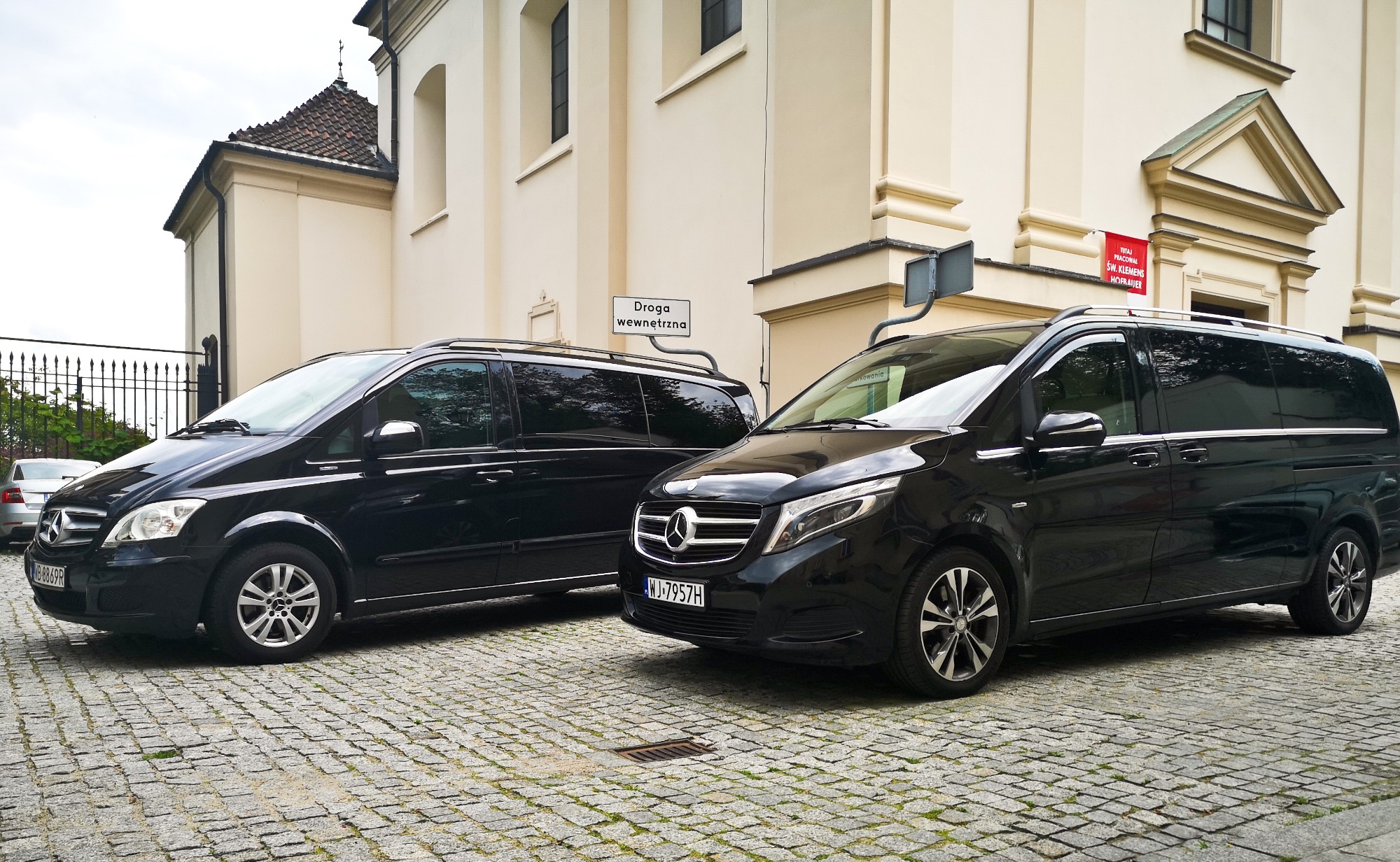 Dwa czarne busy Mercedes-Benz V-klasa zaparkowane na brukowanej ulicy przed budynkiem z beżową elewacją i znakiem drogowym.