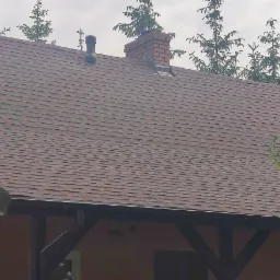 Dach pokryty brązowym gontem bitumicznym, widoczny komin z cegły, rynna metalowa, fragment ściany budynku i otaczająca zieleń.