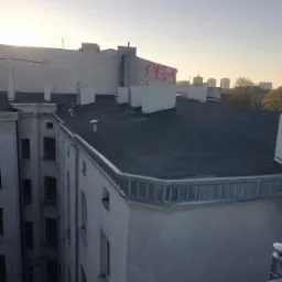 Widok z góry na płaski dach budynku wielorodzinnego z obróbkami blacharskimi, w tle panorama miasta i zachodzące słońce.