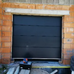 Czarna segmentowa brama garażowa w trakcie montażu w surowym stanie, widoczne cegły i narzędzia instalacyjne.