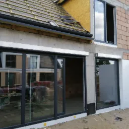 Montaż okien i drzwi przesuwnych w surowym budynku. Widoczne okna z roletami zewnętrznymi i duże, przeszklone drzwi przesuwne z czarnymi ramami.