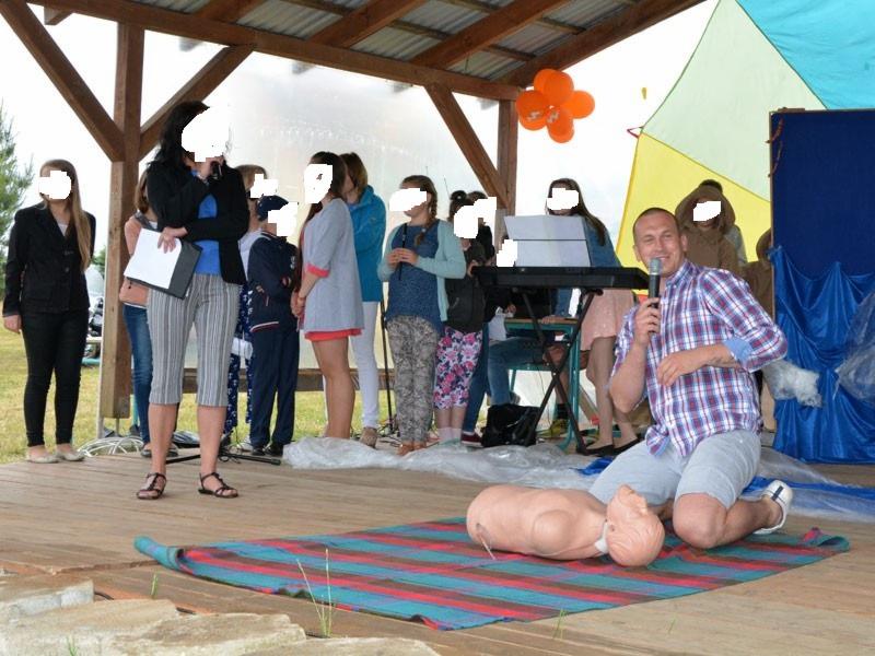 Instruktor demonstruje resuscytację krążeniowo-oddechową na fantomie przed grupą osób na scenie, w tle widoczny keyboard i dekoracje.
