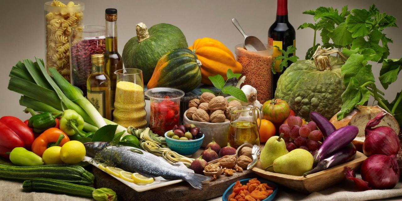 Bogato zaaranżowany stół z różnorodnymi produktami spożywczymi: ryba, warzywa (dynie, papryka, bakłażany, cebula), owoce (gruszki, winogrona, figi), orzechy, oliwa, makaron i przyprawy w słoikach.