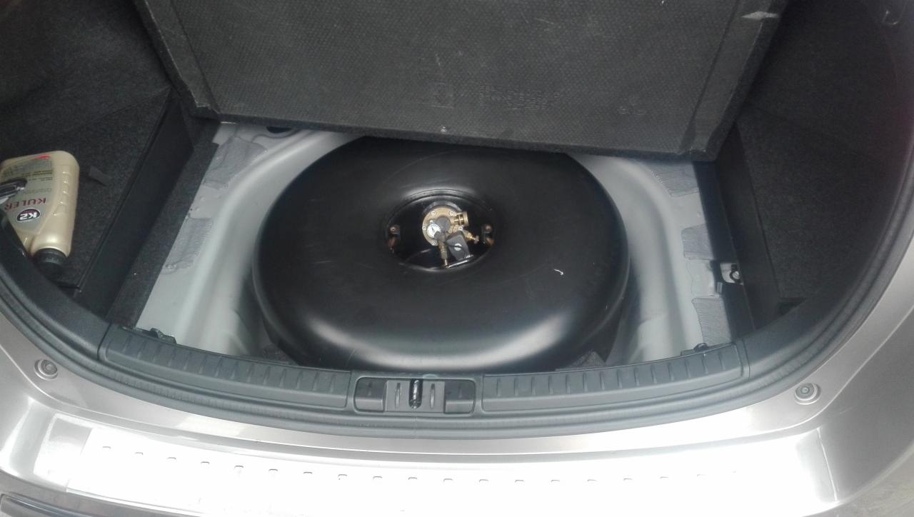 Toroidalny zbiornik na gaz LPG zamontowany w bagażniku samochodu osobowego, widoczny od góry, obok butelka płynu chłodniczego.