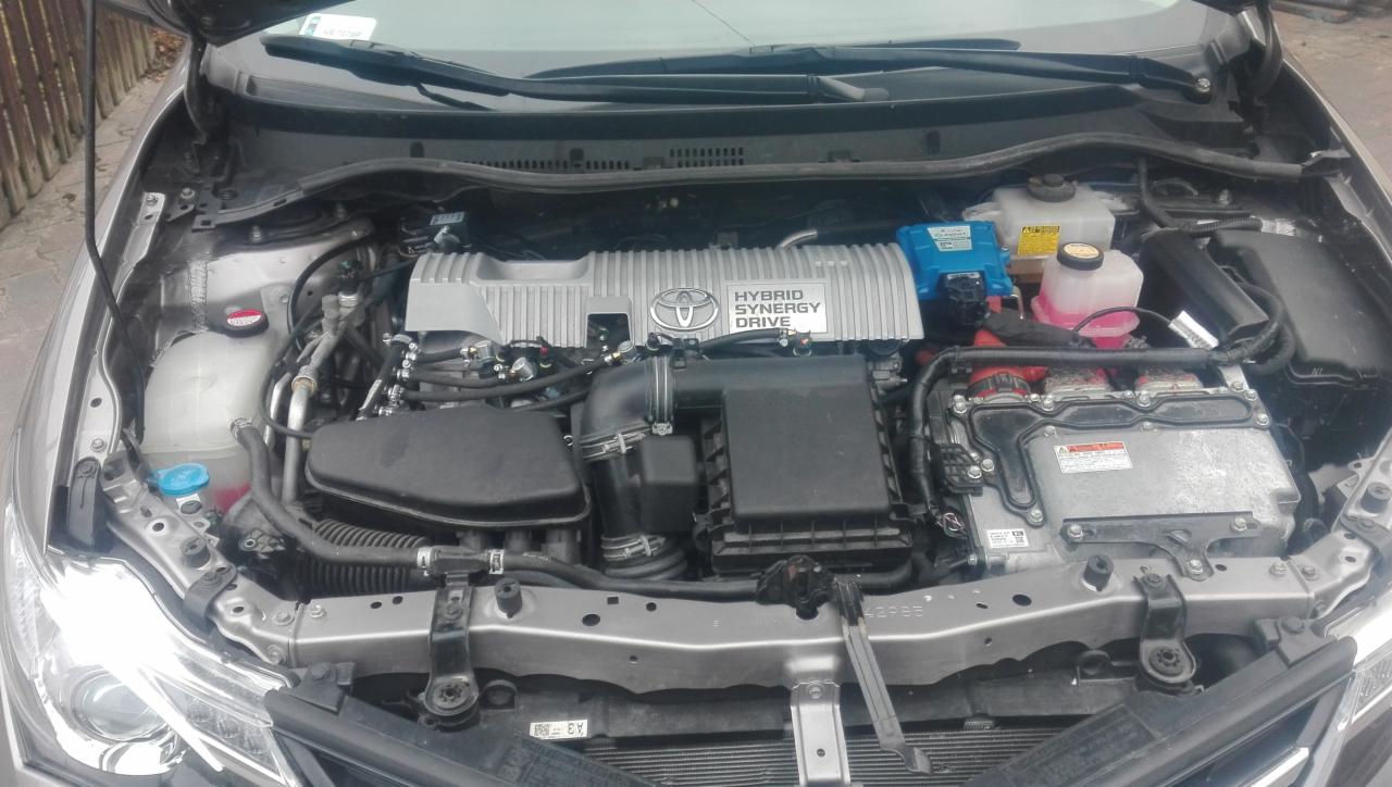 Otwarta maska samochodu hybrydowego, widoczny silnik z napisem HYBRID SYNERGY DRIVE i elementami instalacji elektrycznej, zbiorniczki płynów eksploatacyjnych.