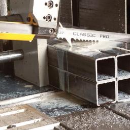 Pragmatec - Automatyczna piła taśmowa przecina wiązkę kwadratowych profili stalowych, chłodziwo spływa po ciętym materiale.
