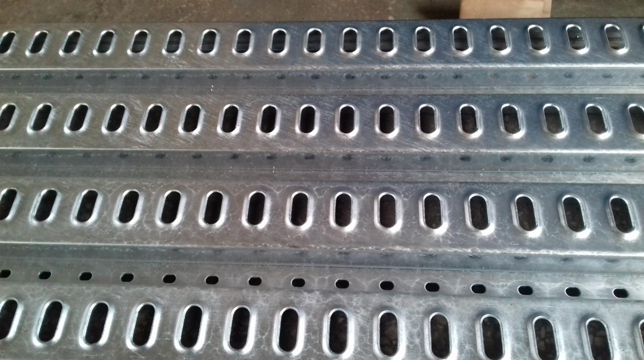 Metalowe profile z regularnymi otworami o owalnym kształcie, ułożone jeden na drugim, widoczne ślady obróbki.