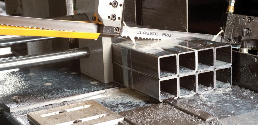 Automatyczna piła taśmowa przecina wiązkę kwadratowych profili stalowych, chłodziwo spływa po ciętym materiale.
