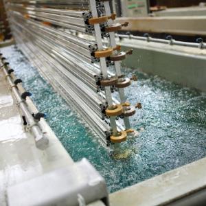 Metalowe profile zanurzone w roztworze podczas obróbki, widoczne mocowania i powierzchnia cieczy z refleksami świetlnymi.
