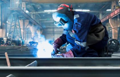 Pracownik w kombinezonie ochronnym i masce spawalniczej łączy metalowe elementy, iskrzące światło w hali produkcyjnej z suwnicą w tle.