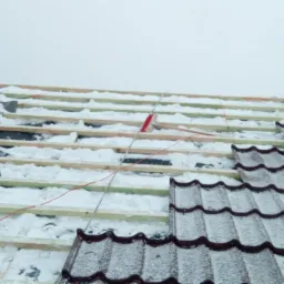 Fragment dachu w trakcie prac, widoczne krokwie pokryte śniegiem, częściowo ułożone brązowe dachówki i ceglany komin w tle.