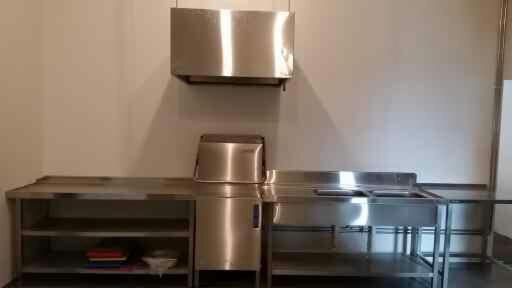 Profesjonalna kuchnia ze stali nierdzewnej z półkami, okapem i zlewem, przygotowana do czyszczenia, z widocznymi śladami użytkowania.