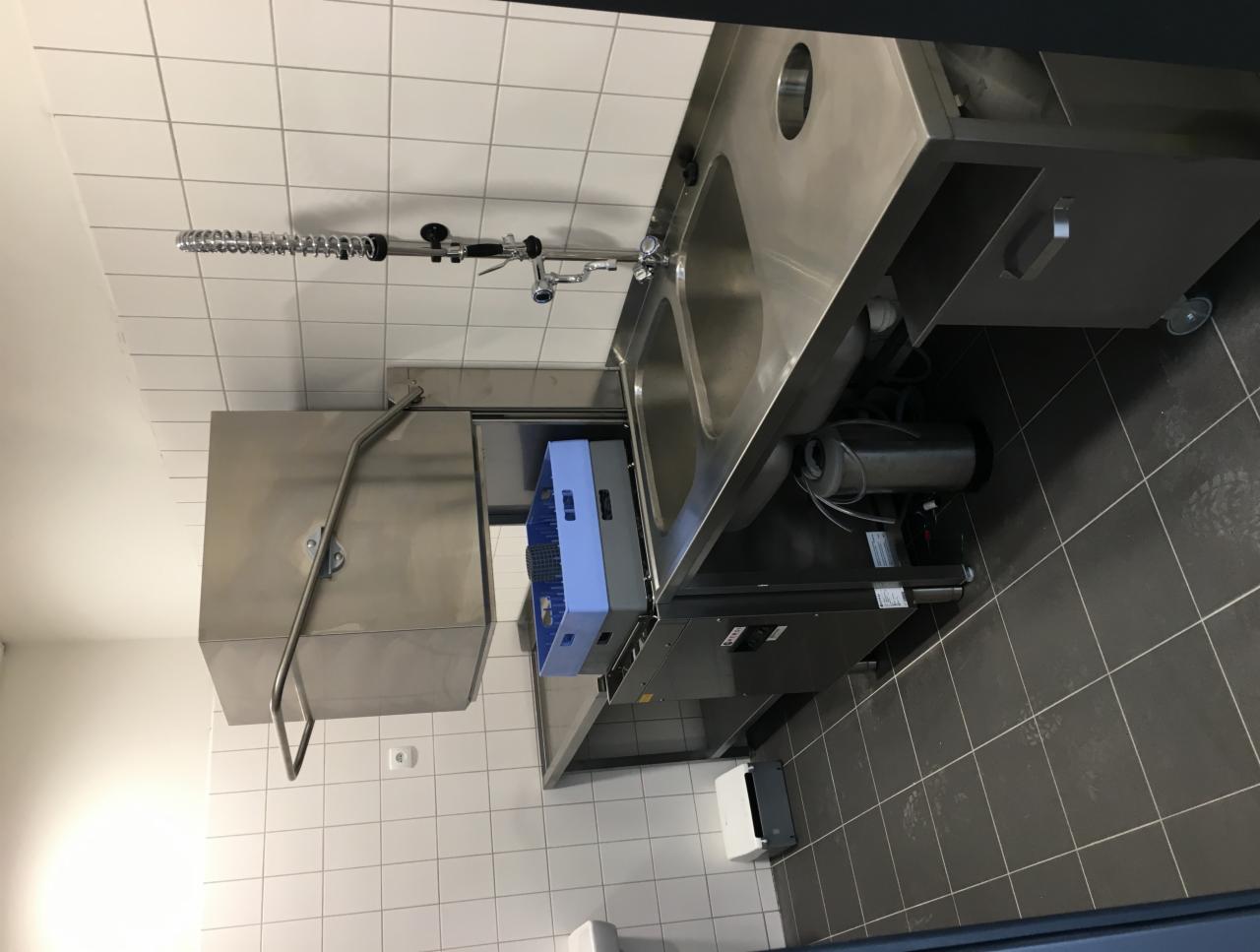 Profesjonalna zmywarka do naczyń ze stali nierdzewnej z koszem na naczynia wewnątrz, widoczna w pomieszczeniu z białymi kafelkami na ścianach i ciemnymi kafelkami na podłodze.