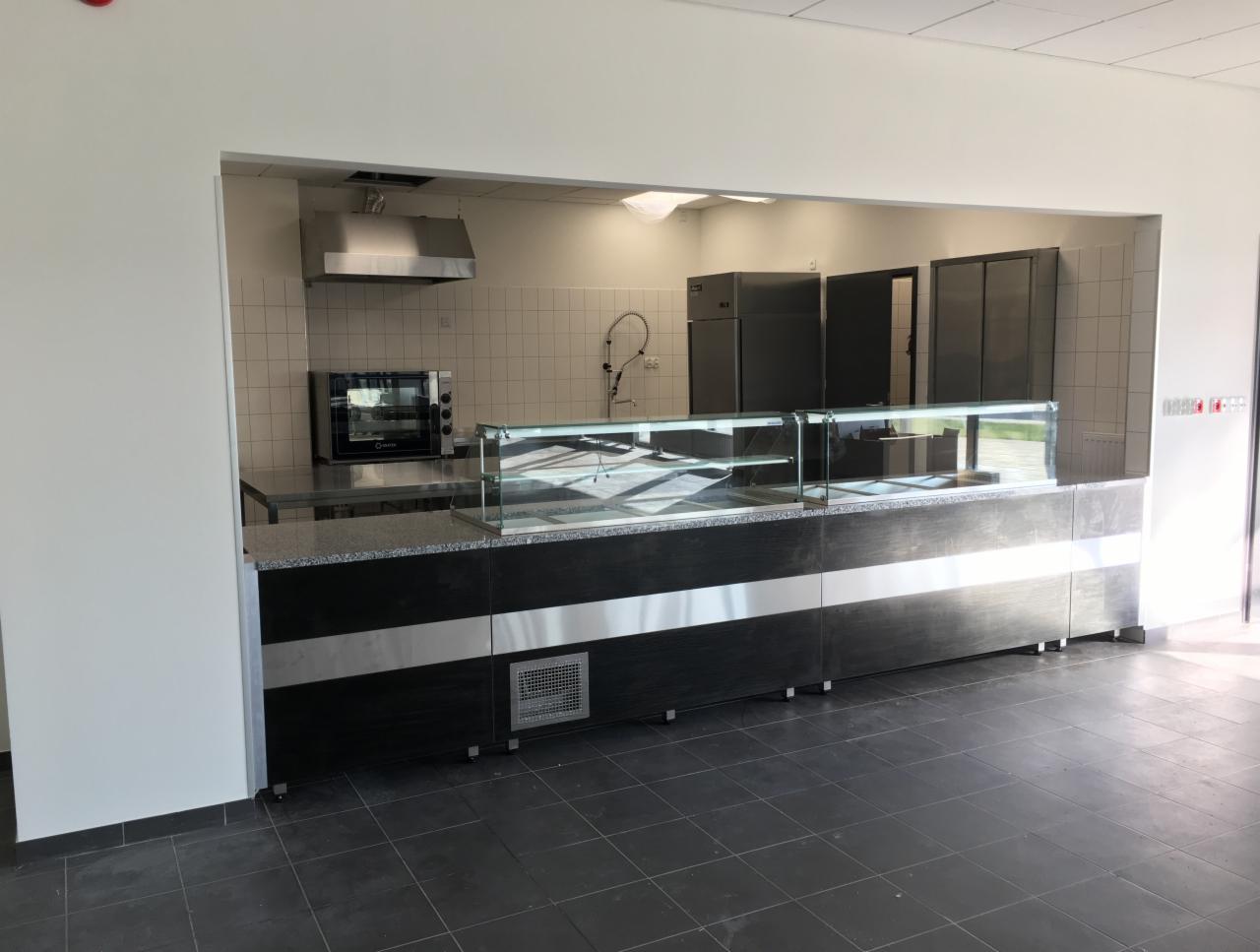 Wnętrze kuchni przemysłowej z widocznym blatem roboczym, witryną chłodniczą, piecem konwekcyjnym i okapem, całość utrzymana w stonowanej kolorystyce.