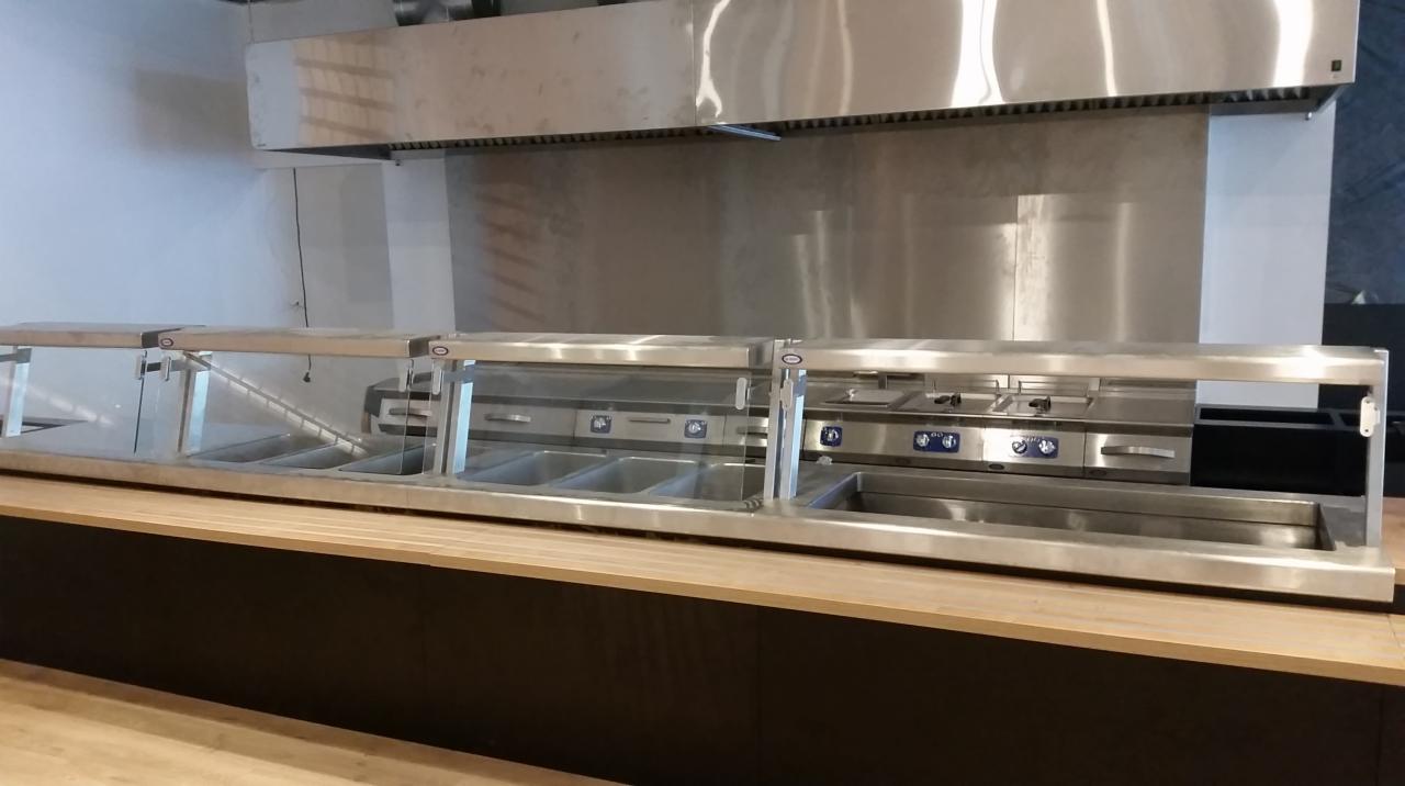 Profesjonalna kuchnia z nierdzewnymi urządzeniami do utrzymywania ciepła potraw, widoczny okap i blat roboczy.