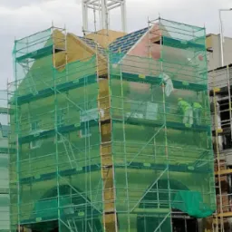 Budynek w trakcie ocieplania, widoczne rusztowania z siatką ochronną, warstwa izolacyjna i pracownicy w kombinezonach ochronnych.