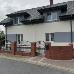 Dwukondygnacyjny dom z szarą dachówką i elewacją w dwóch odcieniach szarości, ogrodzony ceglanym murkiem z metalowymi przęsłami, widoczne rusztowanie przy ścianie szczytowej.