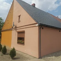 Ocieplony budynek z elewacją w odcieniach beżu i żółci, z oknem z żaluzjami i kwiatami, widoczna antena satelitarna i trzy małe tuje przy ścianie.