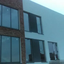 Nowoczesny budynek dwukondygnacyjny z elewacją w dwóch kolorach: biały tynk i czerwona cegła, duże okna z ciemnymi ramami, widok z dołu w pochmurny dzień.