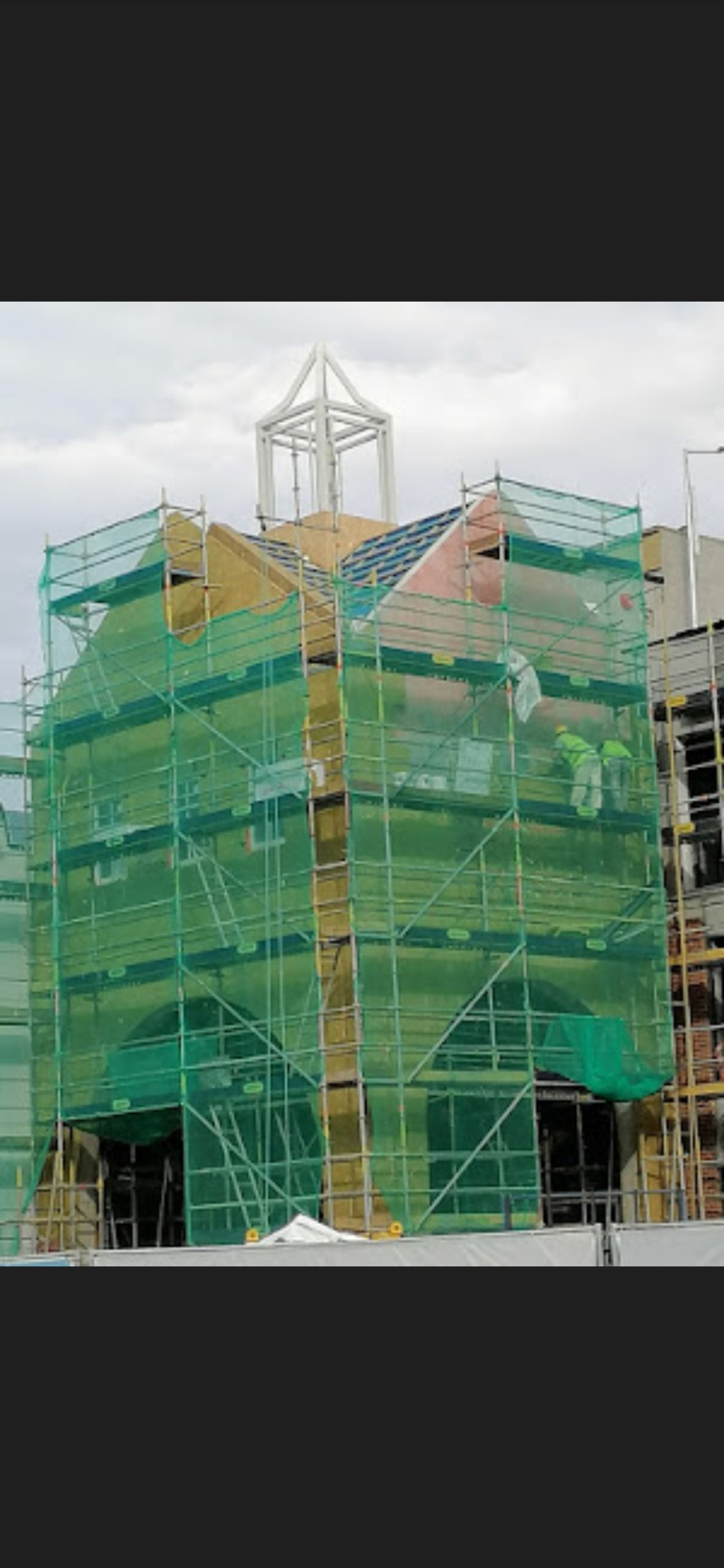 Budynek w trakcie ocieplania, widoczne rusztowania z siatką ochronną, warstwa izolacyjna i pracownicy w kombinezonach ochronnych.