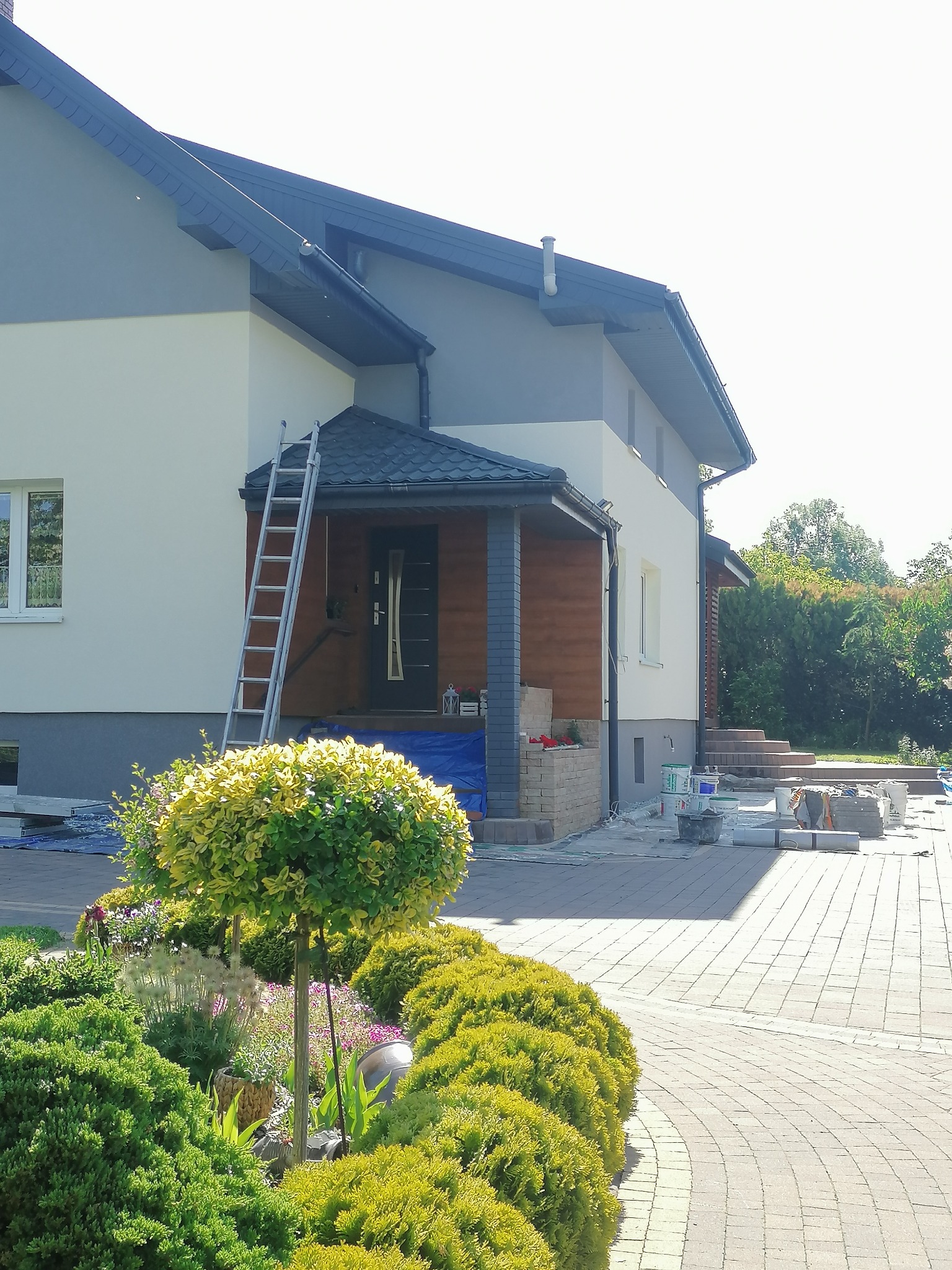 Dwukondygnacyjny dom jednorodzinny z szarą elewacją górnej części i jasną dolnej, z drewnianą werandą i aluminiową drabiną opartą o ścianę, widoczne prace wykończeniowe i materiały budowlane...