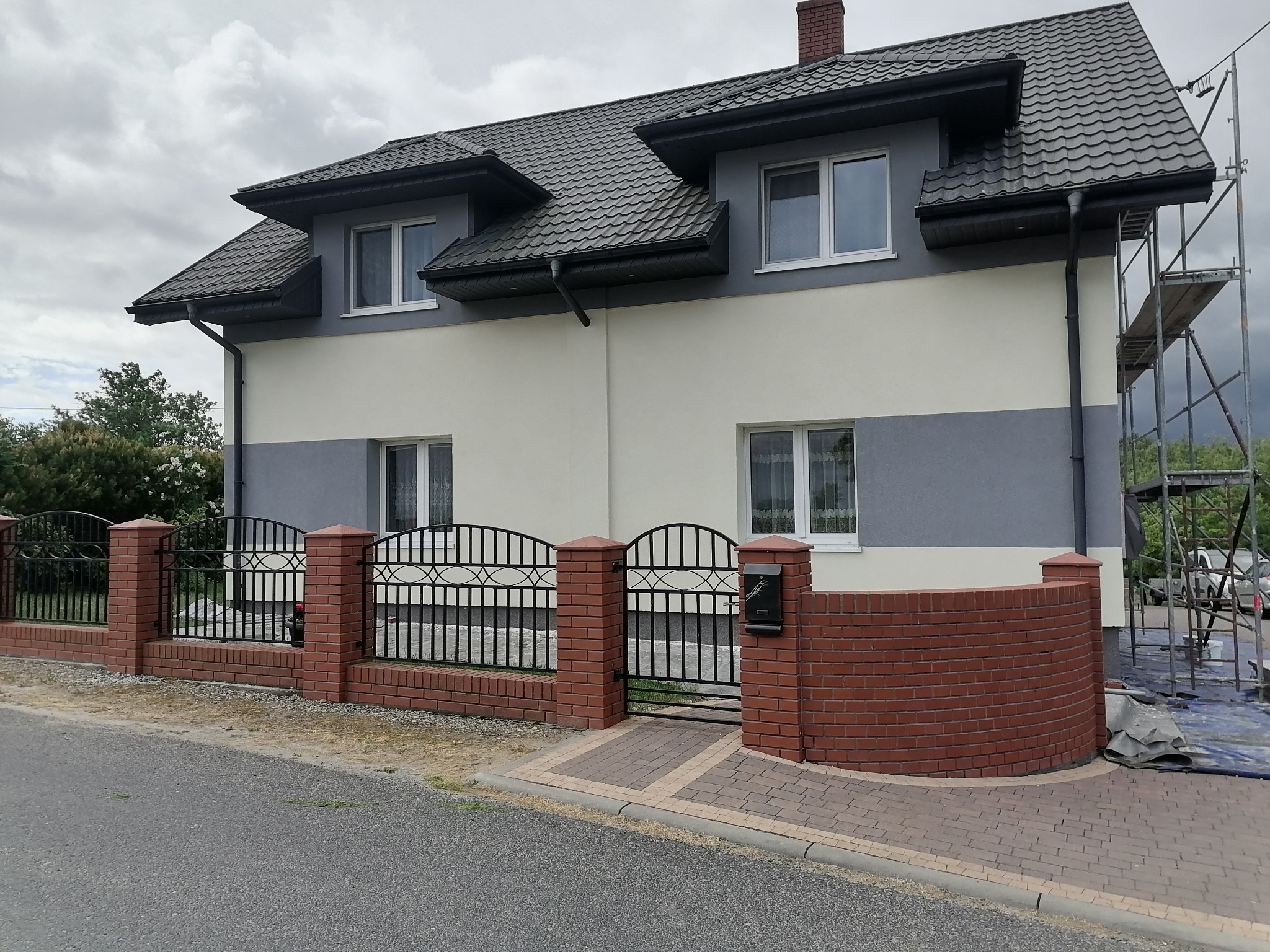 Dwukondygnacyjny dom z szarą dachówką i elewacją w dwóch odcieniach szarości, ogrodzony ceglanym murkiem z metalowymi przęsłami, widoczne rusztowanie przy ścianie szczytowej.