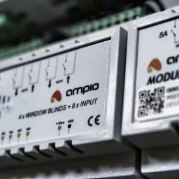 Szczegółowe ujęcie modułów sterujących systemem inteligentnego domu marki Ampio, widoczne schematy połączeń i oznaczenia CE, QR kod, moduł do sterowania roletami okiennymi.