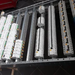 Rozdzielnica elektryczna w trakcie montażu, widoczne moduły zabezpieczeń z podłączonymi przewodami w kolorach zielonym, pomarańczowym i czerwonym, zamontowane na szynach DIN wewnątrz metalowej ramy.