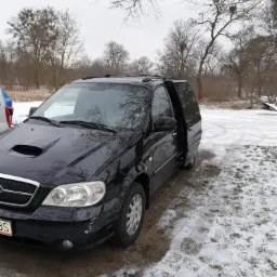 Czarny minivan Kia Carnival z otwartymi drzwiami przesuwnymi, stojący na ziemi pokrytej śniegiem i trawą, zarys drzew w tle.