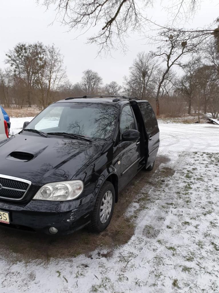 Czarny minivan Kia Carnival z otwartymi drzwiami przesuwnymi, stojący na ziemi pokrytej śniegiem i trawą, zarys drzew w tle.