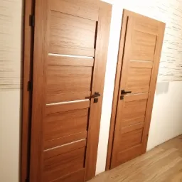 Dwa skrzydła drzwiowe z drewnianą okleiną i poziomymi przeszkleniami, widoczne czarne klamki i zawiasy, zamontowane w nowym budownictwie z panelami podłogowymi.