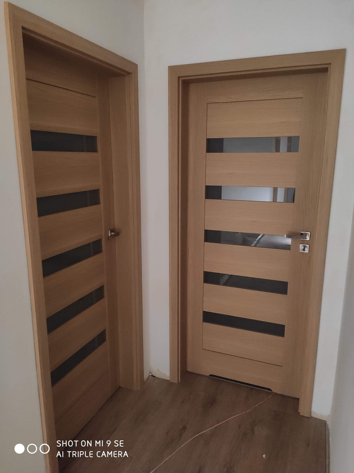 Dwa skrzydła drzwiowe z jasnego drewna z poziomymi, ciemnymi przeszkleniami, zamontowane w białej ścianie, widoczne na tle brązowej podłogi.