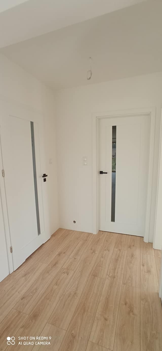 Białe drzwi wewnętrzne z czarną klamką i szybą, widoczne na tle jasnych ścian i podłogi z paneli, w nowym mieszkaniu po remoncie.