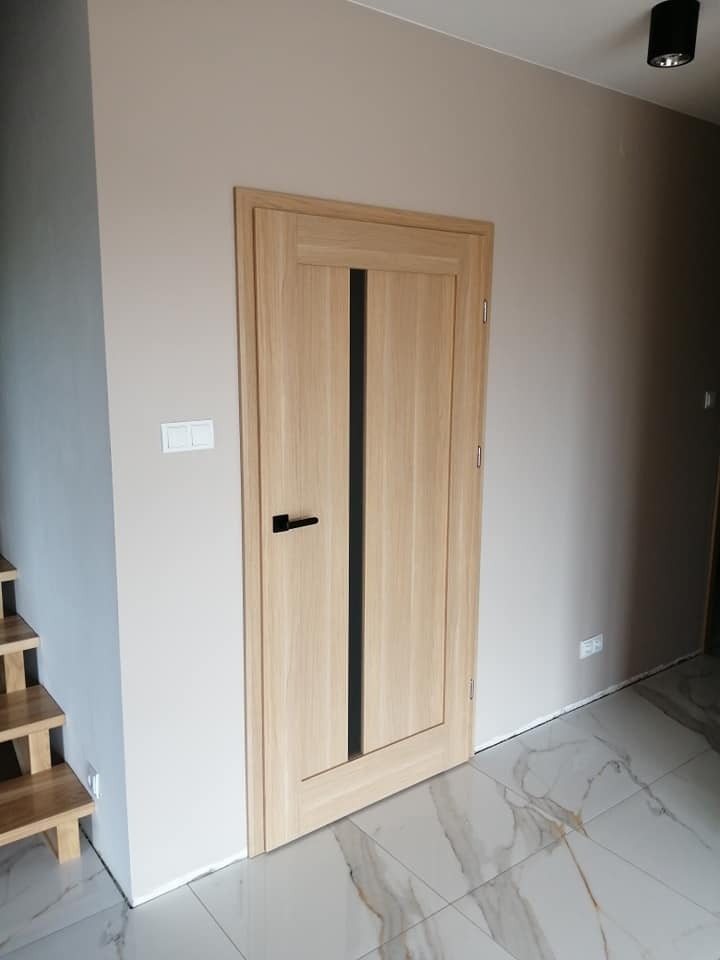 Jasne, drewniane drzwi z czarnym, pionowym panelem i czarną klamką, zamontowane w beżowej ścianie, widoczne fragmenty drewnianych schodów i marmurowej podłogi.