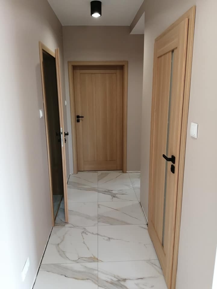 Korytarz z trzema drewnianymi drzwiami o różnej konstrukcji i czarnymi klamkami, wykończony marmurowymi płytkami podłogowymi i oświetleniem punktowym.