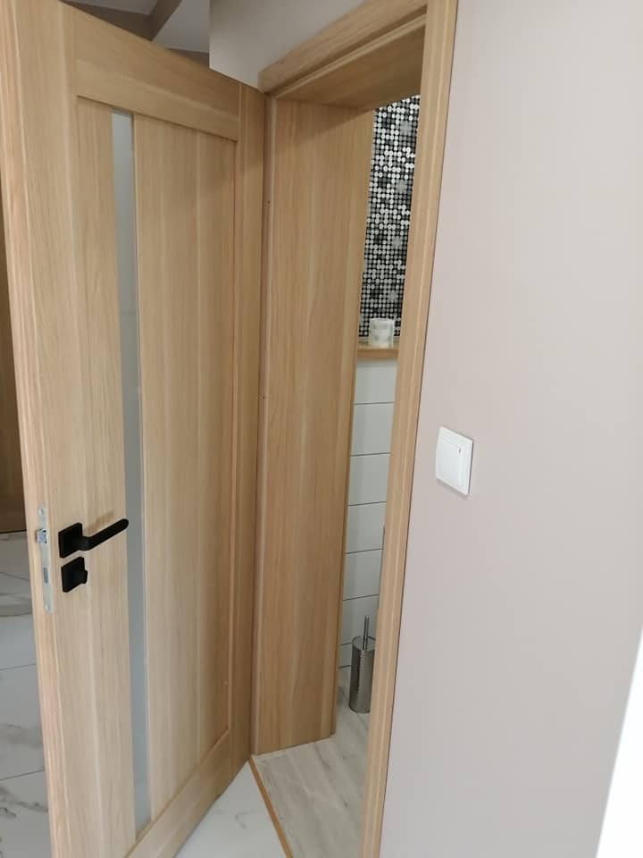 Jasne drewniane drzwi z matowym przeszkleniem i czarną klamką, widok na łazienkę z mozaiką w tle.