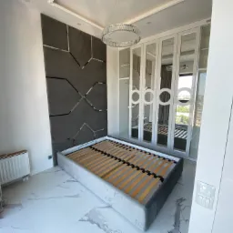 Szary tapicerowany stelaż łóżka z drewnianymi listwami w nowoczesnej sypialni z dekoracyjną ścianą i lustrzaną szafą.