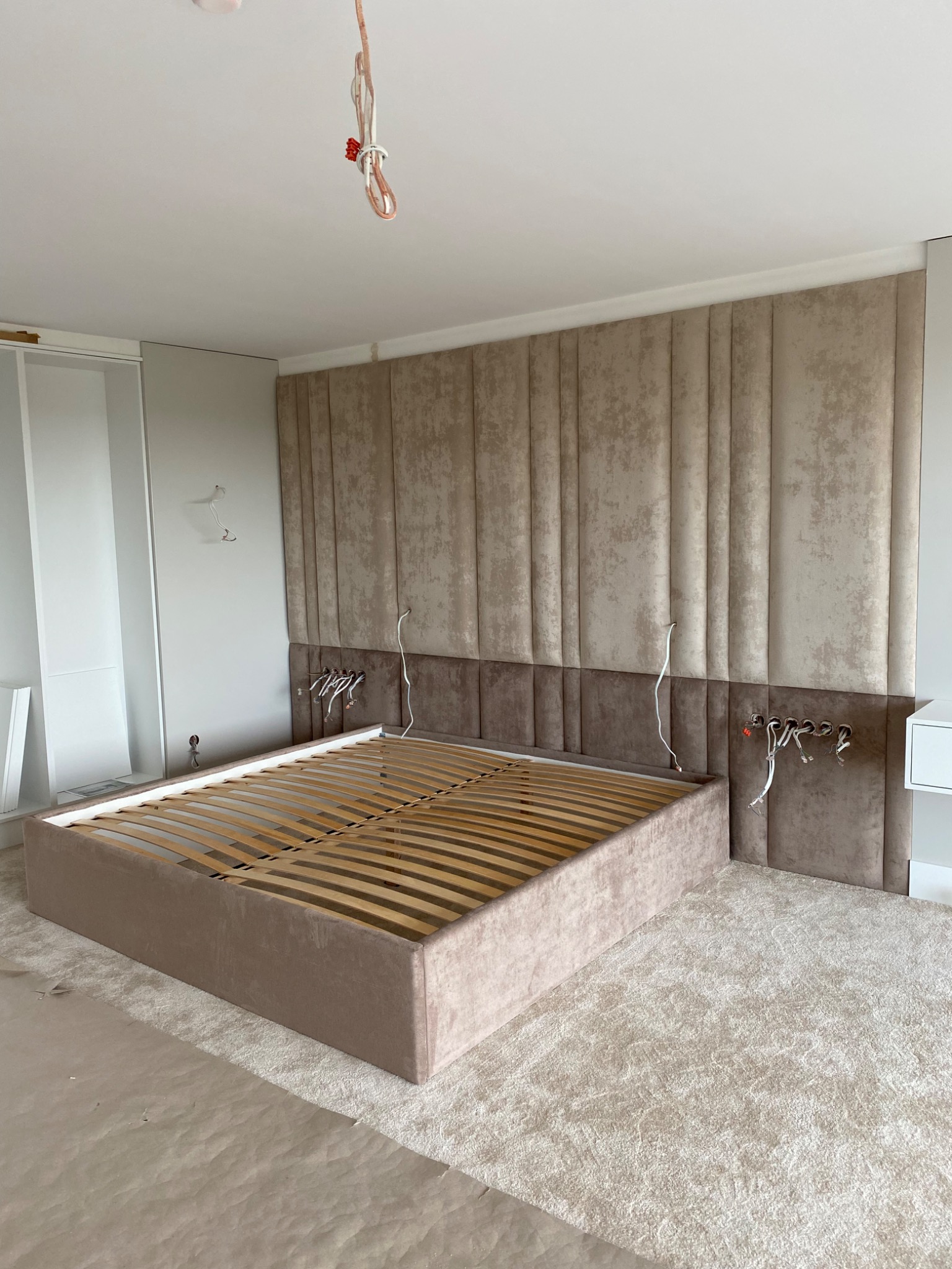 Beżowe łóżko z drewnianym stelażem i tapicerowanym wezgłowiem w trakcie montażu, widoczne kable elektryczne wystające ze ściany, w tle jasna wykładzina.