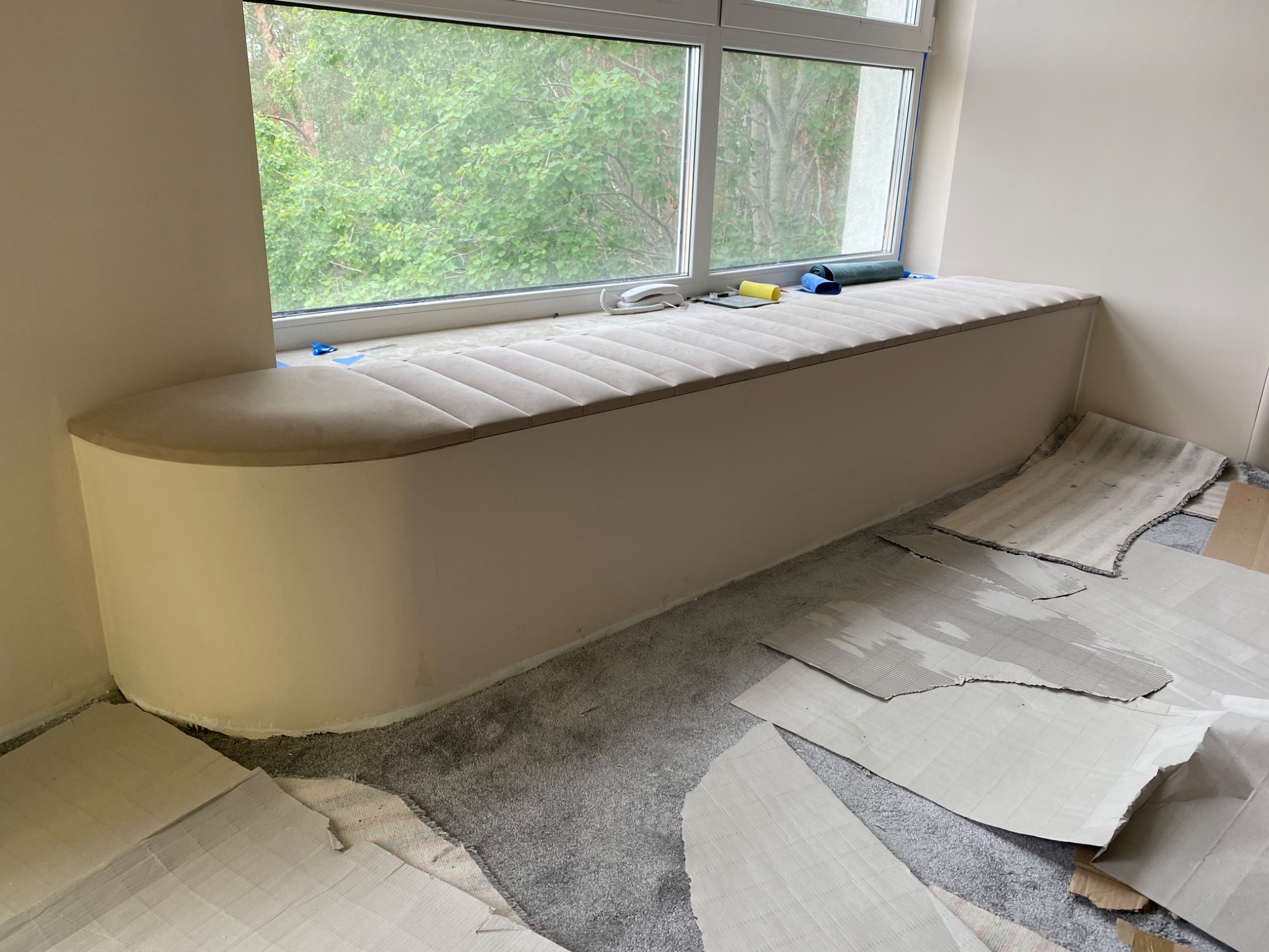 Wnęka okienna z siedziskiem tapicerowanym w odcieniach beżu, widoczne narzędzia i materiały tapicerskie, podłoga zabezpieczona kartonem, okno z widokiem na zieleń.