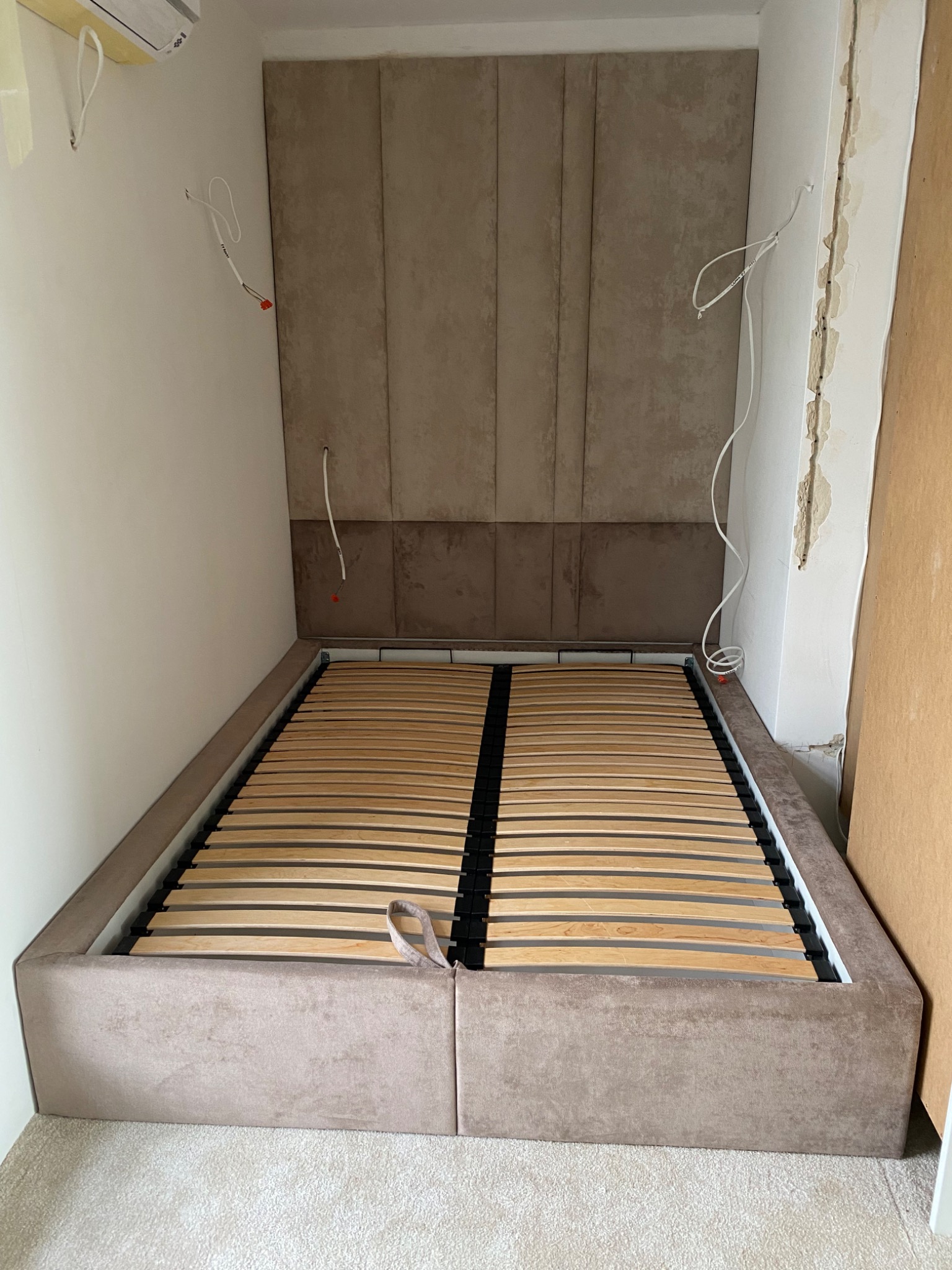 Rama łóżka z drewnianym stelażem i wezgłowiem obitym beżową tkaniną, widoczne przewody elektryczne wystające ze ściany, sugerujące prace wykończeniowe.