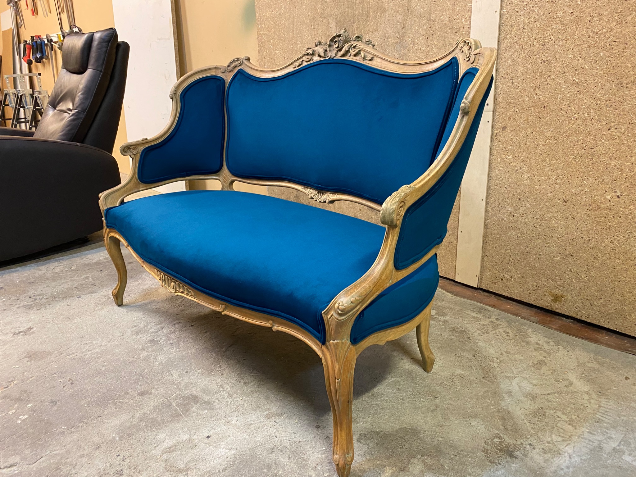 Odrestaurowana sofa w stylu Ludwika Filipa, obita turkusowym welurem, z widocznymi detalami rzeźbionego, jasnego drewna. W tle warsztat z narzędziami i czarnym fotelem.
