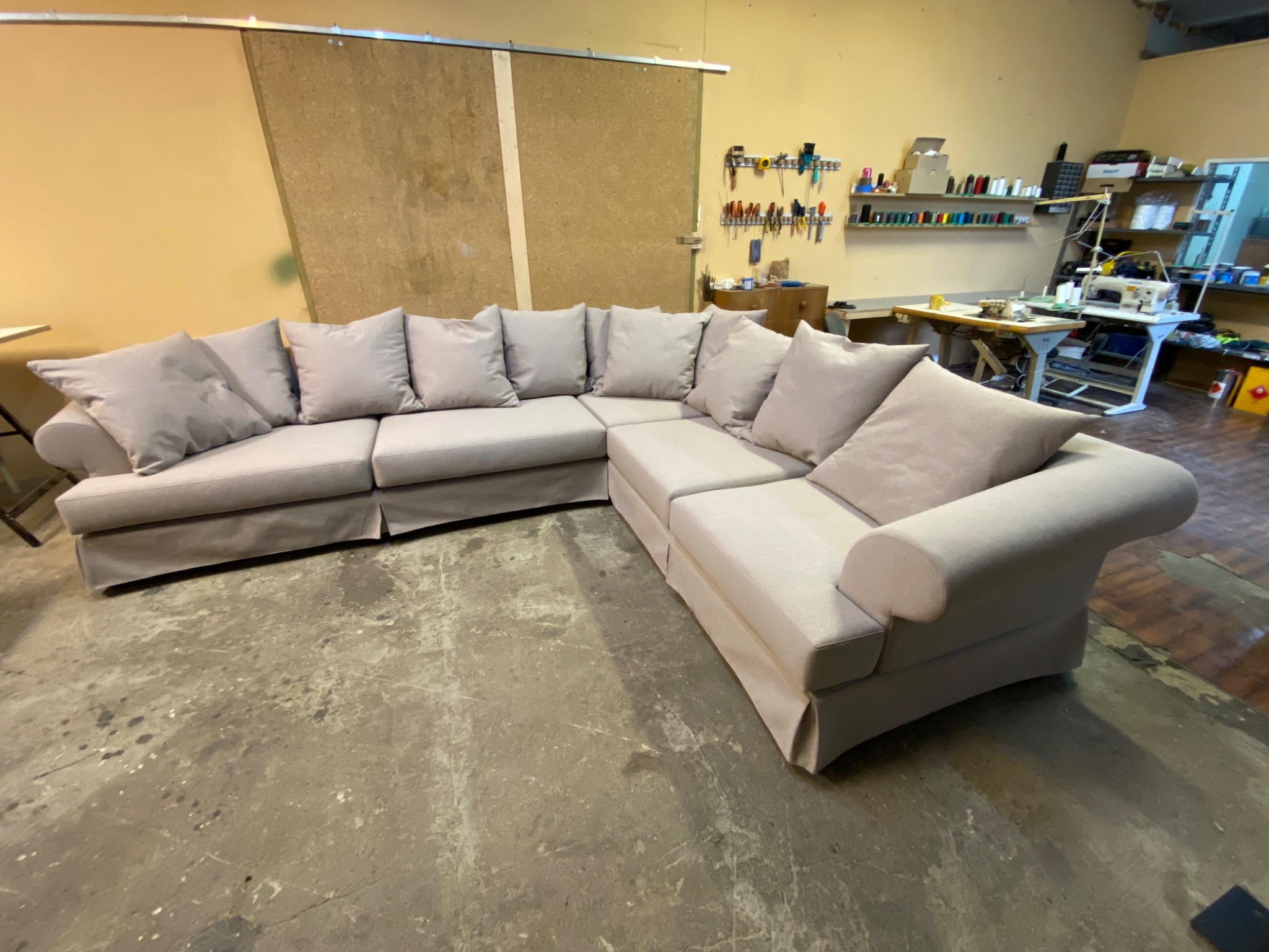 Narożnik z jasną tapicerką i poduszkami w warsztacie, widoczne narzędzia i maszyny do szycia w tle.