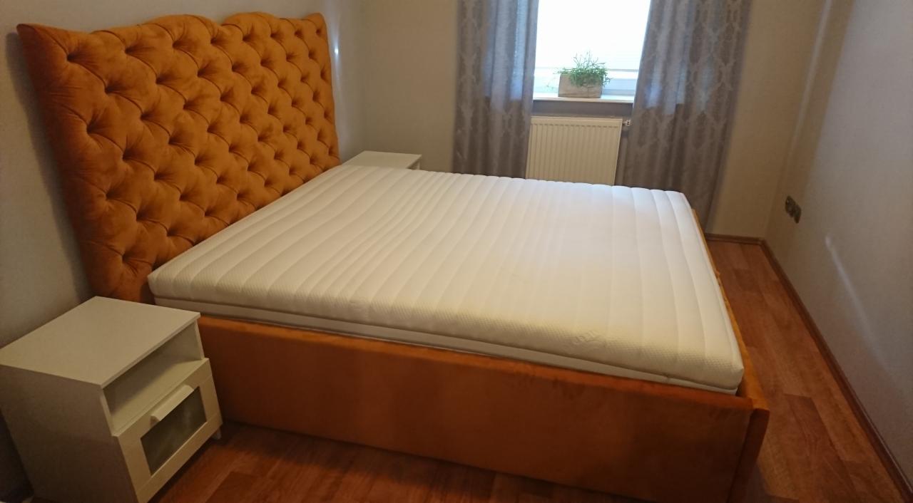 Pomarańczowe łóżko tapicerowane z pikowanym zagłówkiem i białą szafką nocną, widok z boku na tle okna z zasłonami.