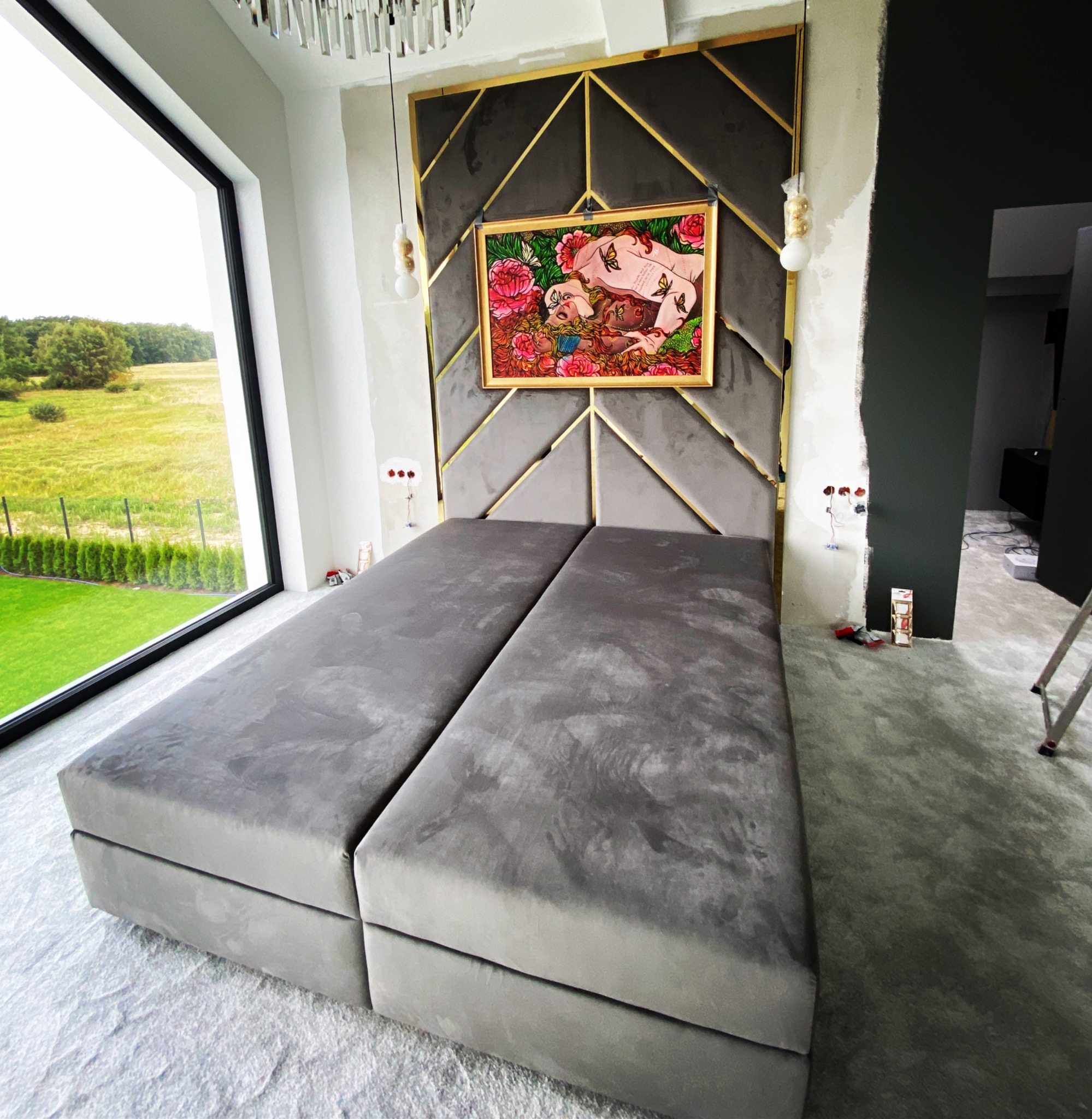 Dwa szare, tapicerowane łóżka kontynentalne z wezgłowiem w geometryczne wzory i złotymi akcentami, wiszące lampy i obraz w złotej ramie, widok na zielony krajobraz przez duże okno, pokój w trakcie...